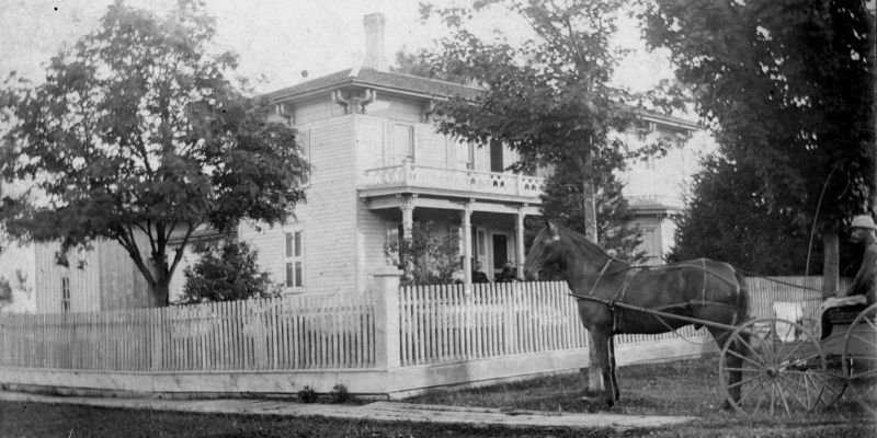 Black and white image of white two-story wood-sided house with horse standing outside the fence