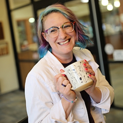 Woman in a white shirt smiling and holding a coffee mug in front of glass windows