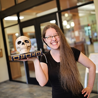Brunette woman in a black shirt and glasses holding up a book and a skull