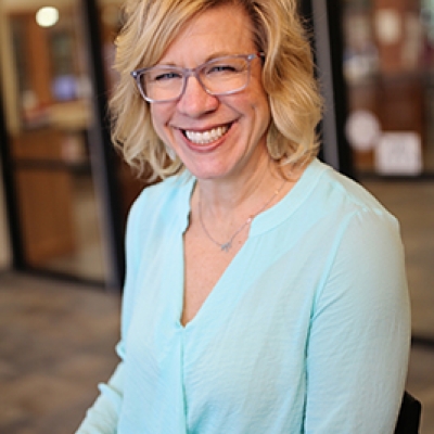 Blonde woman in a blue shirt and glasses with glass windows in the background