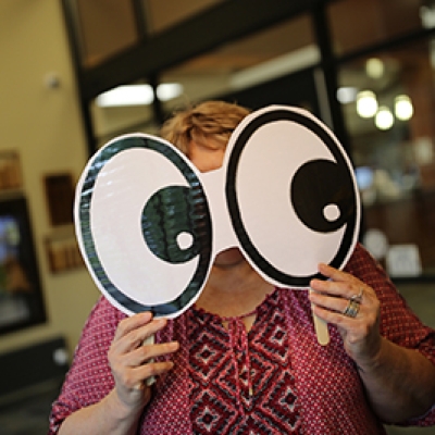 Lady in a pink shirt holding up large paper eyes in front of her face with glass windows in the background