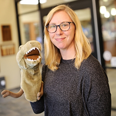 Blonde woman with a black shirt and glasses wearing a dinosaur arm puppet