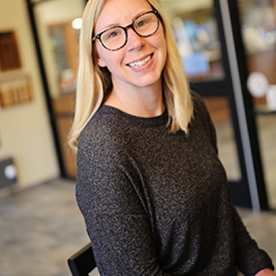 Blonde lady in a black shirt and glasses sitting on a high chair with glass windows behind her